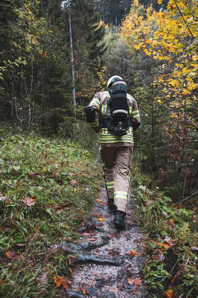 Feuerwehrteilnehmer mit Atemschutzgerät steigt auf einem schmalen, nassen Waldpfad über Wurzeln.