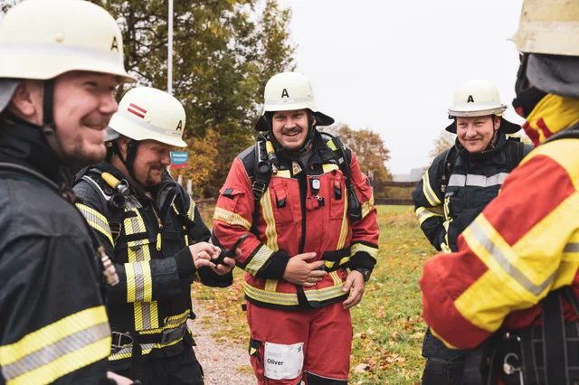 Lachende Gruppe in roter und schwarzer Einsatzkleidung mit hellen Helmen.