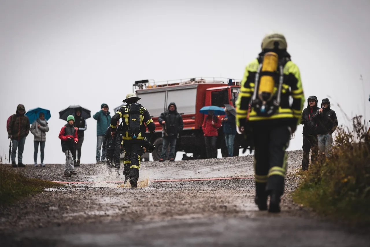 Feuerwehrleute laufen durch Pfützen zur Zielmarkierung vor Zuschauern mit Regenschirmen.