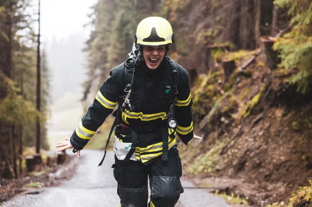 Feuerwehrteilnehmerin mit gelbem Helm lacht mit seitlich ausgestreckten Armen auf der nassen Bergstraße.