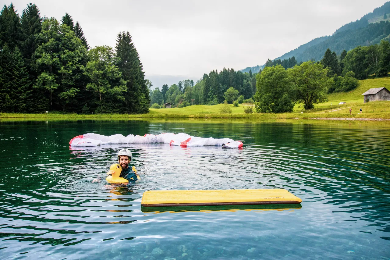 Manuel Nübel schwimmt mit einer gelben Badeente im Teich; sein Gleitschirm treibt hinter ihm auf dem Wasser.