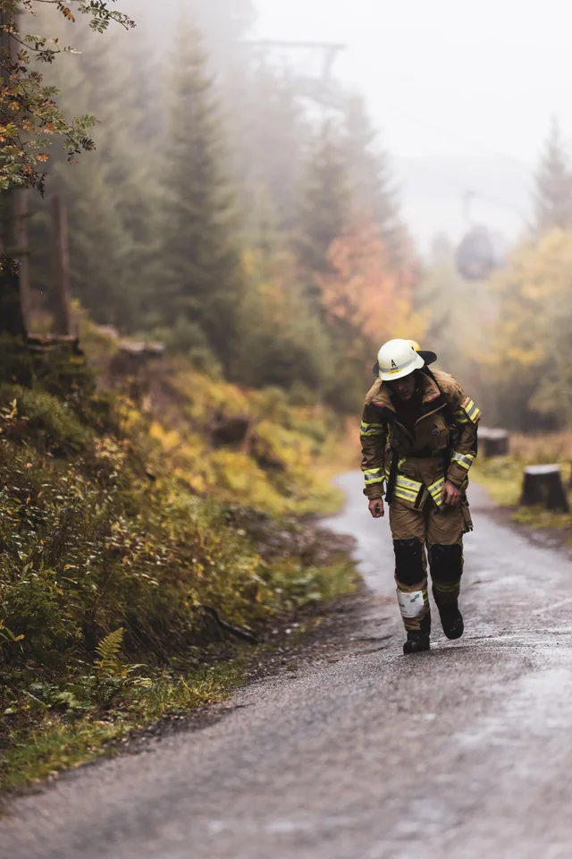 Teilnehmer in beiger Einsatzkleidung steigt vornübergebeugt die nasse Bergstraße hinauf.