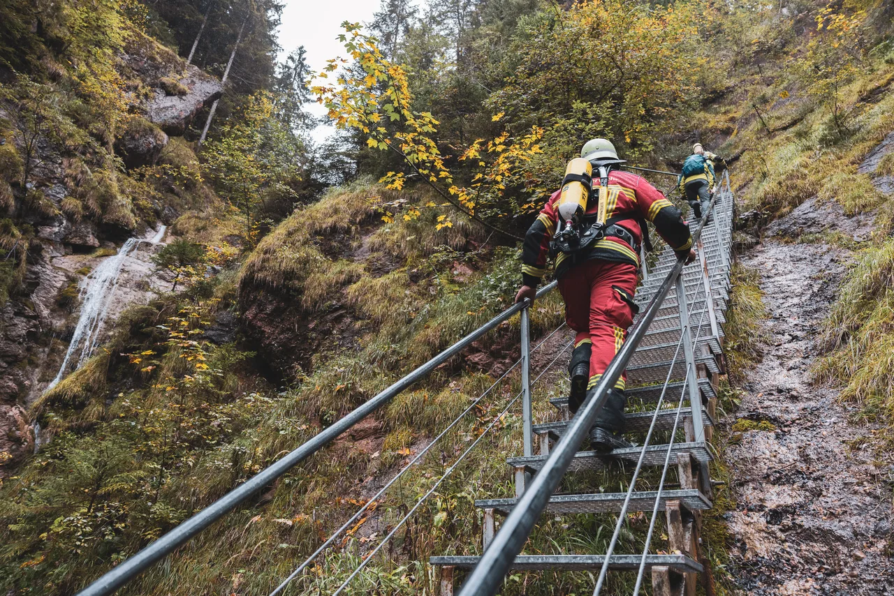 Feuerwehrleute steigen eine steile Metalltreppe neben einem Wasserfall im herbstlichen Wald hinauf.