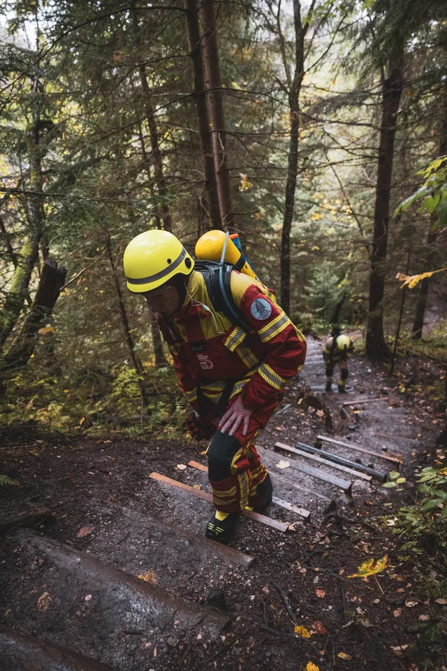 Teilnehmer in roter Einsatzkleidung beugt sich beim Aufstieg über die Waldstufen nach vorn.