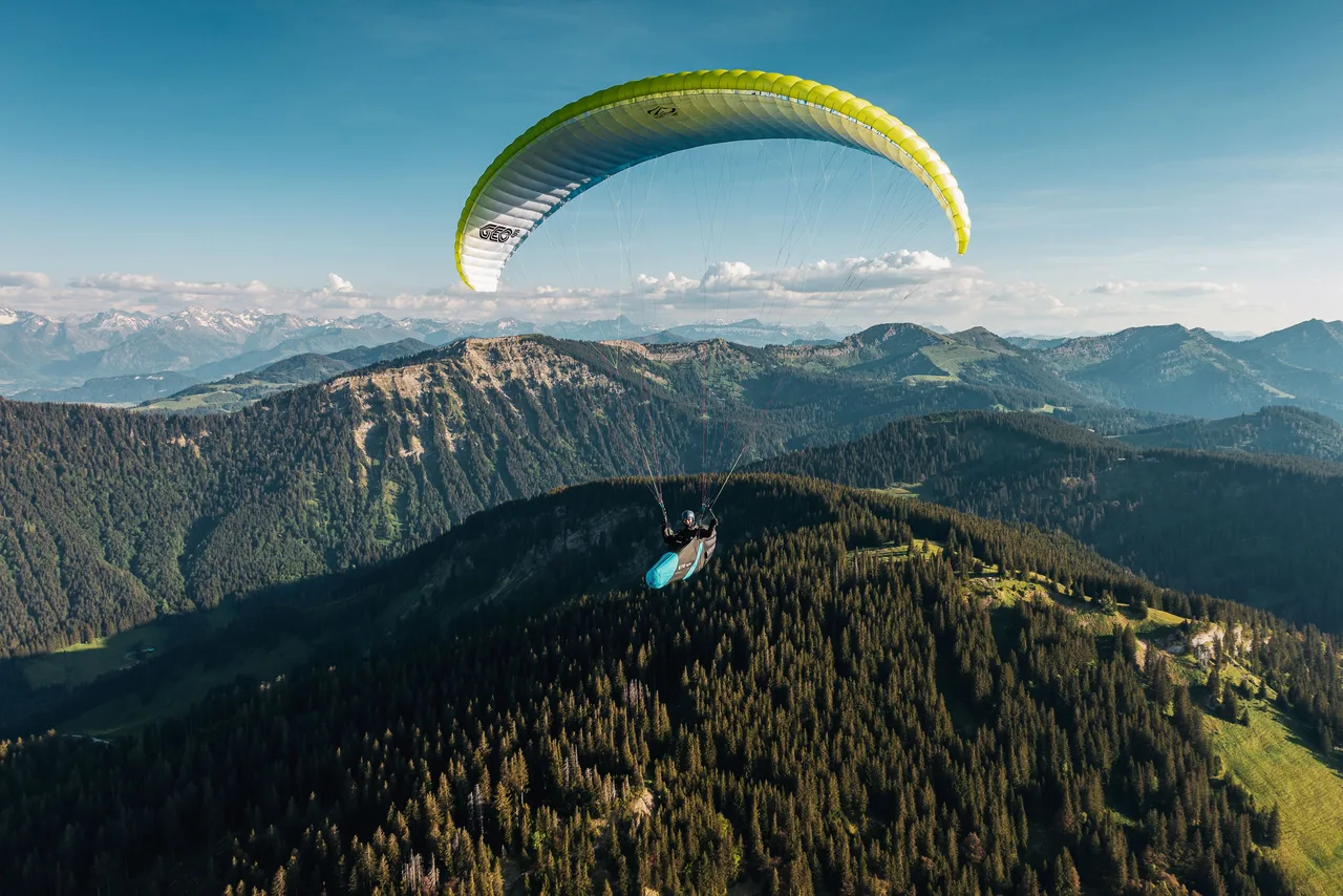 Ein gelbgrüner Gleitschirm mit Pilot fliegt nah über einem bewaldeten Bergrücken vor den Allgäuer Alpen.