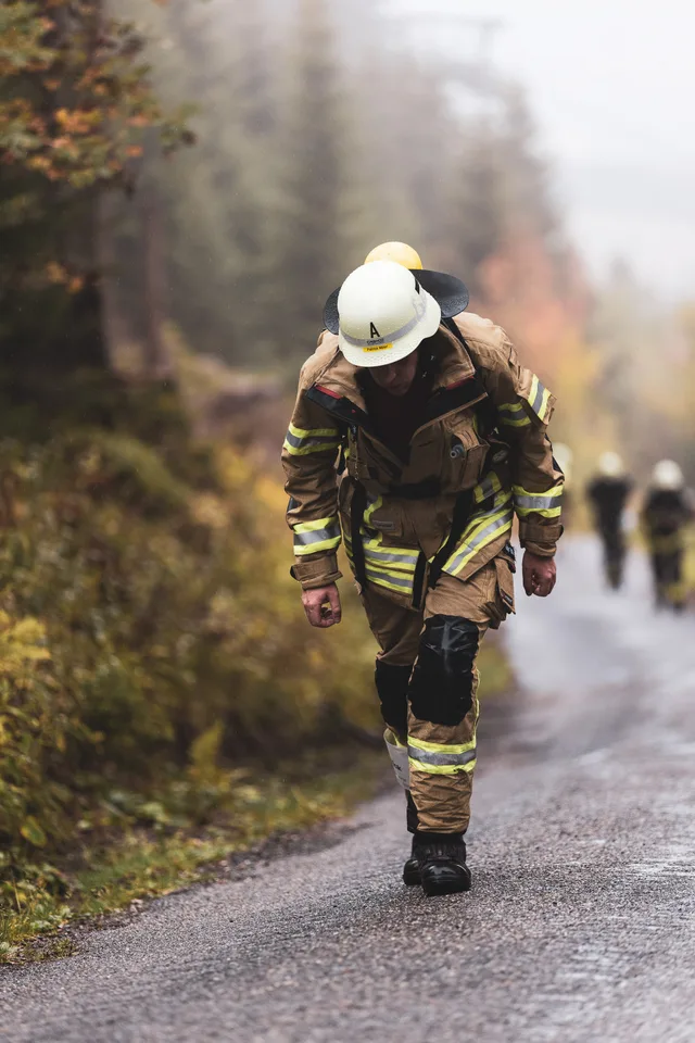 Teilnehmer in beiger Einsatzkleidung steigt vornübergebeugt die nasse Bergstraße hinauf.