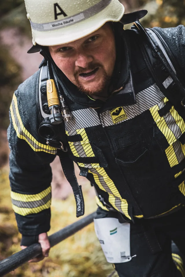 Feuerwehrmann mit regennassem Helm und Atemschutzgerät greift beim Aufstieg an ein Geländer.