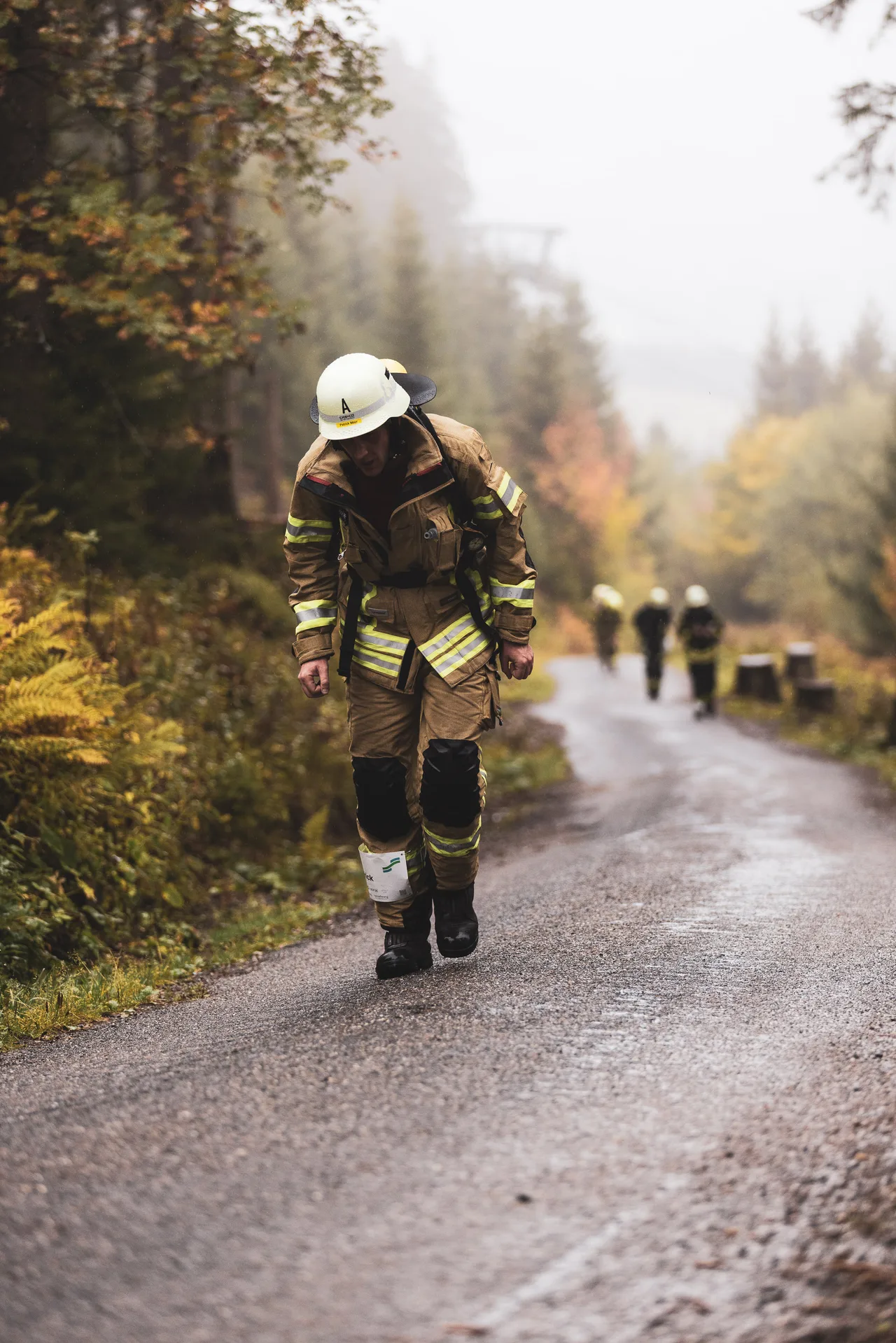 Feuerwehrmann in beiger Schutzkleidung steigt mit gesenktem Kopf eine nasse Straße im Herbstwald hinauf.