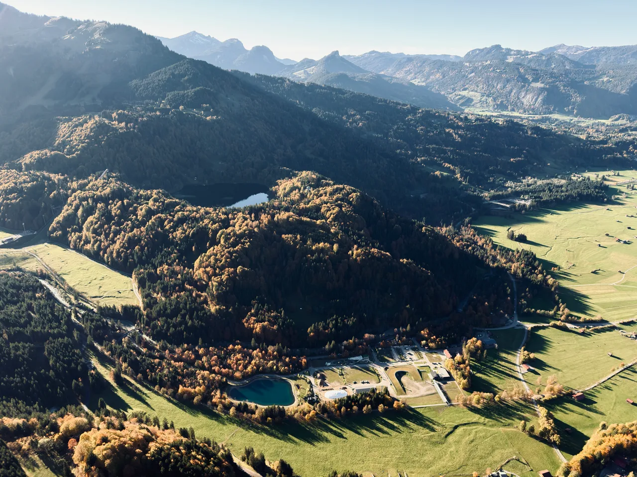 Blick aus der Luft auf den Freibergsee zwischen goldenem Herbstwald und grünen Wiesen bei Oberstdorf