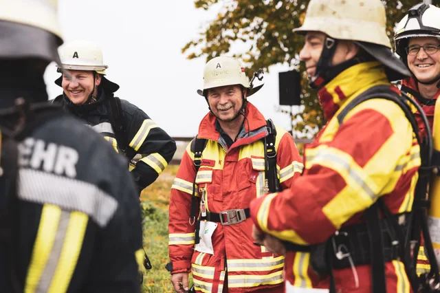 Lachende Gruppe in roter und schwarzer Einsatzkleidung mit hellen Helmen.
