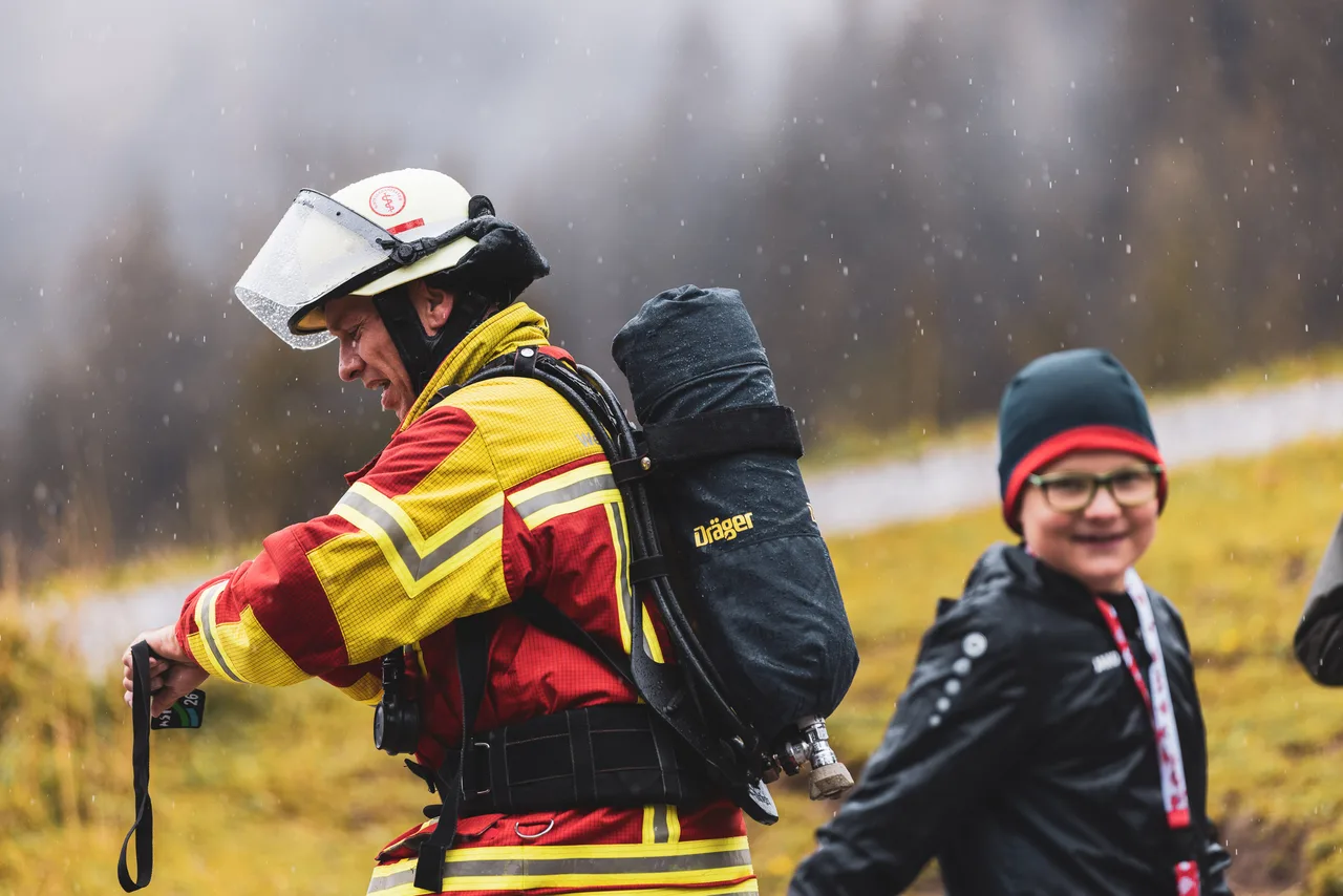 Teilnehmer in rot-gelber Schutzkleidung betrachtet im Regen seine Medaille neben einem Kind.