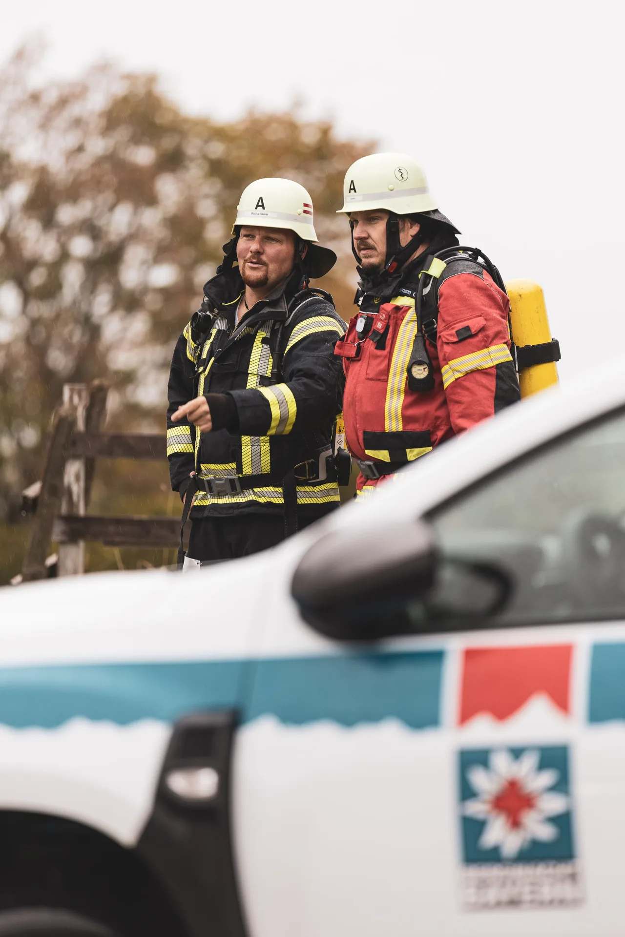 Zwei Feuerwehrleute mit Helm und Atemschutzgerät warten hinter einem Fahrzeug der Bergwacht.