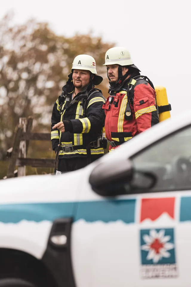 Zwei Feuerwehrleute mit Helm und Atemschutzgerät warten hinter einem Fahrzeug der Bergwacht.