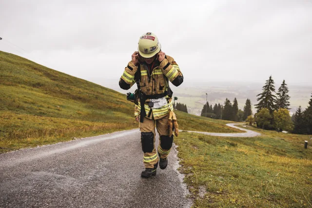Teilnehmer in beiger Einsatzkleidung hält die Tragegurte auf der nassen Bergstraße.