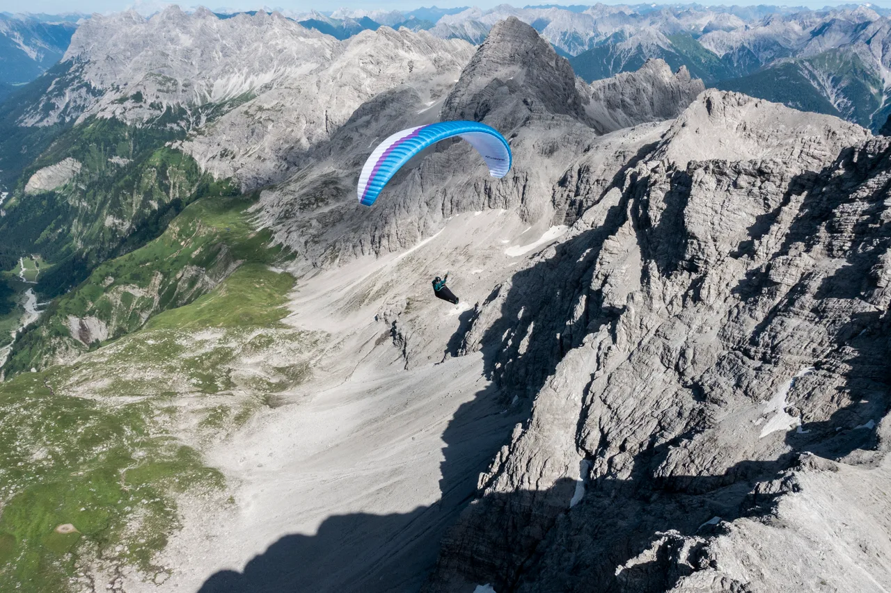 Lara Weinfurtner fliegt unter einem blauen Gleitschirm neben einer steilen Kalkwand über einer hellen Schuttrinne.