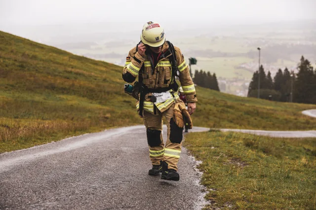 Teilnehmer in beiger Einsatzkleidung hält die Tragegurte auf der nassen Bergstraße.
