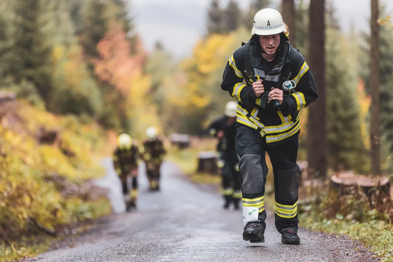 Feuerwehrmann steigt mit Atemschutzgerät und beiden Händen an den Tragegurten eine regennasse Bergstraße hinauf.