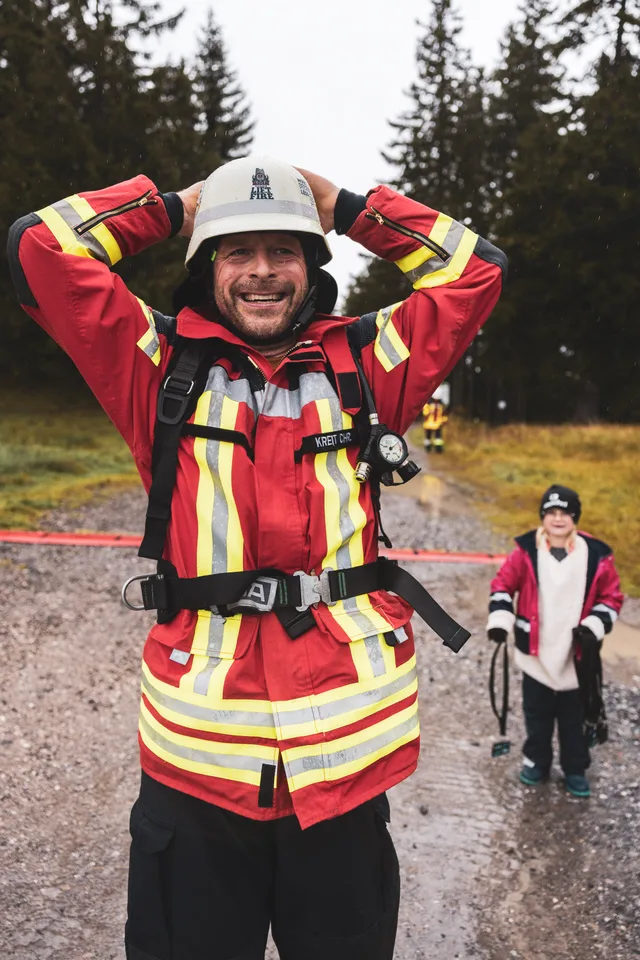 Lachender Feuerwehrmann in roter Schutzkleidung legt beide Hände an seinen Helm.