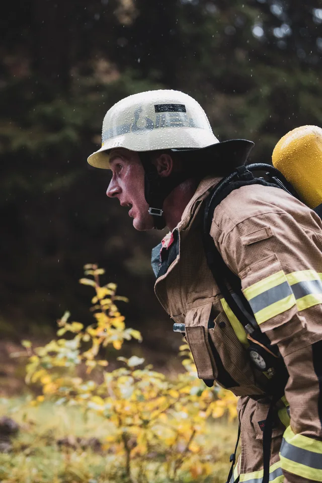 Seitenporträt eines Feuerwehrmanns mit regennassem weißem Helm und gelber Atemluftflasche beim Aufstieg.