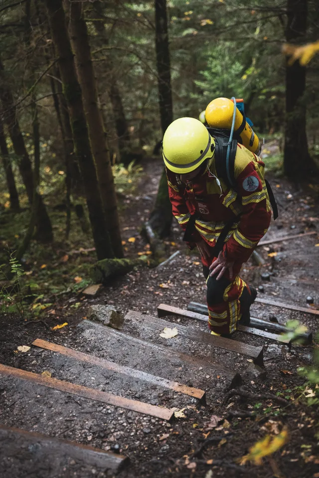 Teilnehmer in roter Einsatzkleidung beugt sich beim Aufstieg über die Waldstufen nach vorn.
