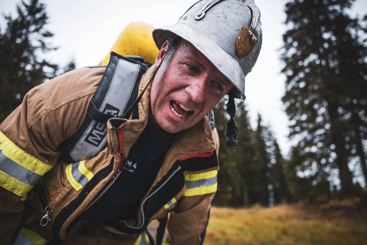 Feuerwehrmann mit silbernem Helm beugt sich mit geöffnetem Mund nach vorn; Tropfen liegen auf seinem Helm.