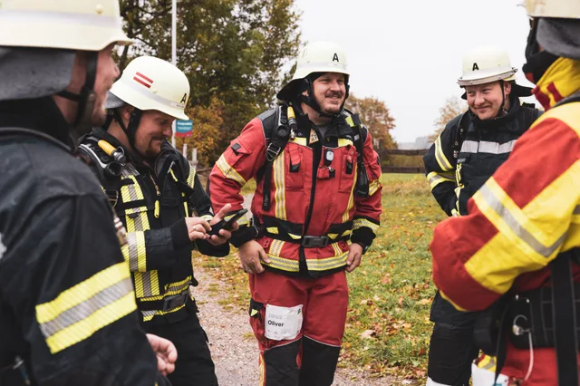 Lachende Gruppe in roter und schwarzer Einsatzkleidung mit hellen Helmen.
