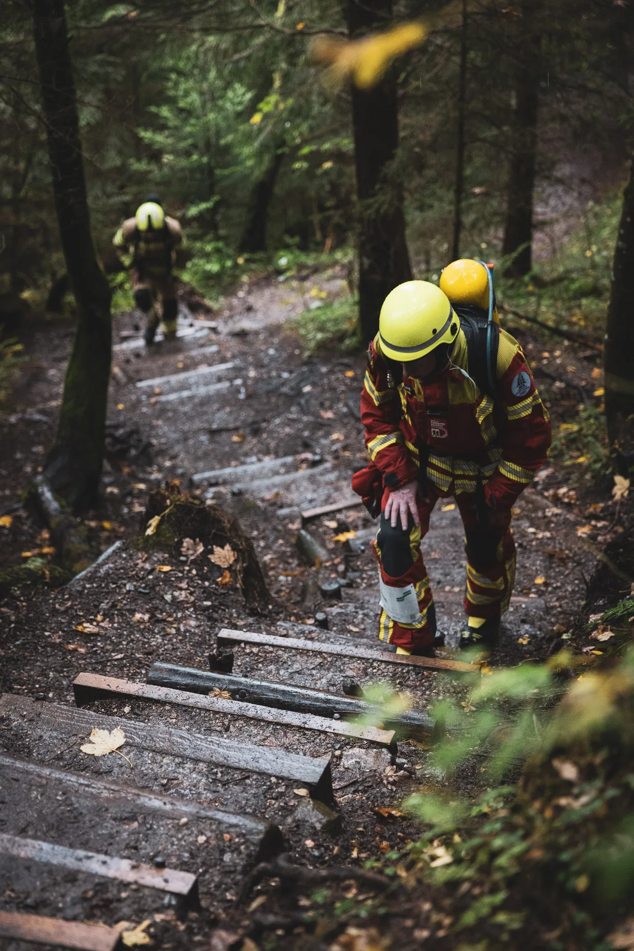 Teilnehmer in roter Schutzkleidung stützt sich beim Aufstieg über nasse Waldstufen auf ein Knie.