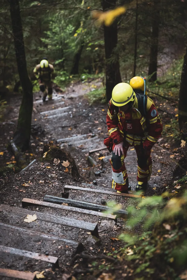 Teilnehmer in roter Schutzkleidung stützt sich beim Aufstieg über nasse Waldstufen auf ein Knie.