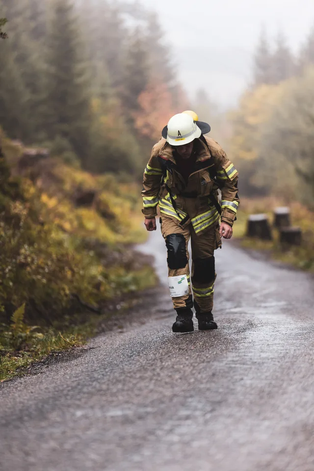 Teilnehmer in beiger Einsatzkleidung steigt vornübergebeugt die nasse Bergstraße hinauf.