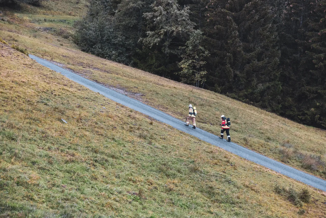 Zwei Feuerwehrleute mit Atemschutzgeräten auf einer steilen Straße zwischen Bergwiese und Wald.
