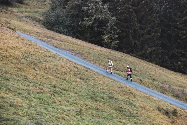 Zwei Feuerwehrleute mit Atemschutzgeräten auf einer steilen Straße zwischen Bergwiese und Wald.