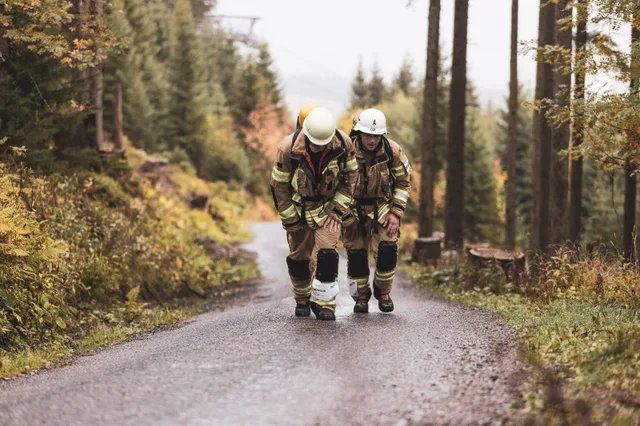 Zwei Teilnehmer in beiger Einsatzkleidung gehen nebeneinander die nasse Bergstraße hinauf.