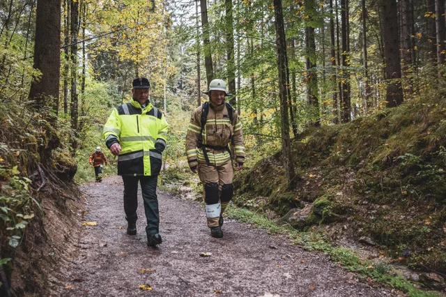 Teilnehmer in beiger Einsatzkleidung neben einem Helfer im Wald.