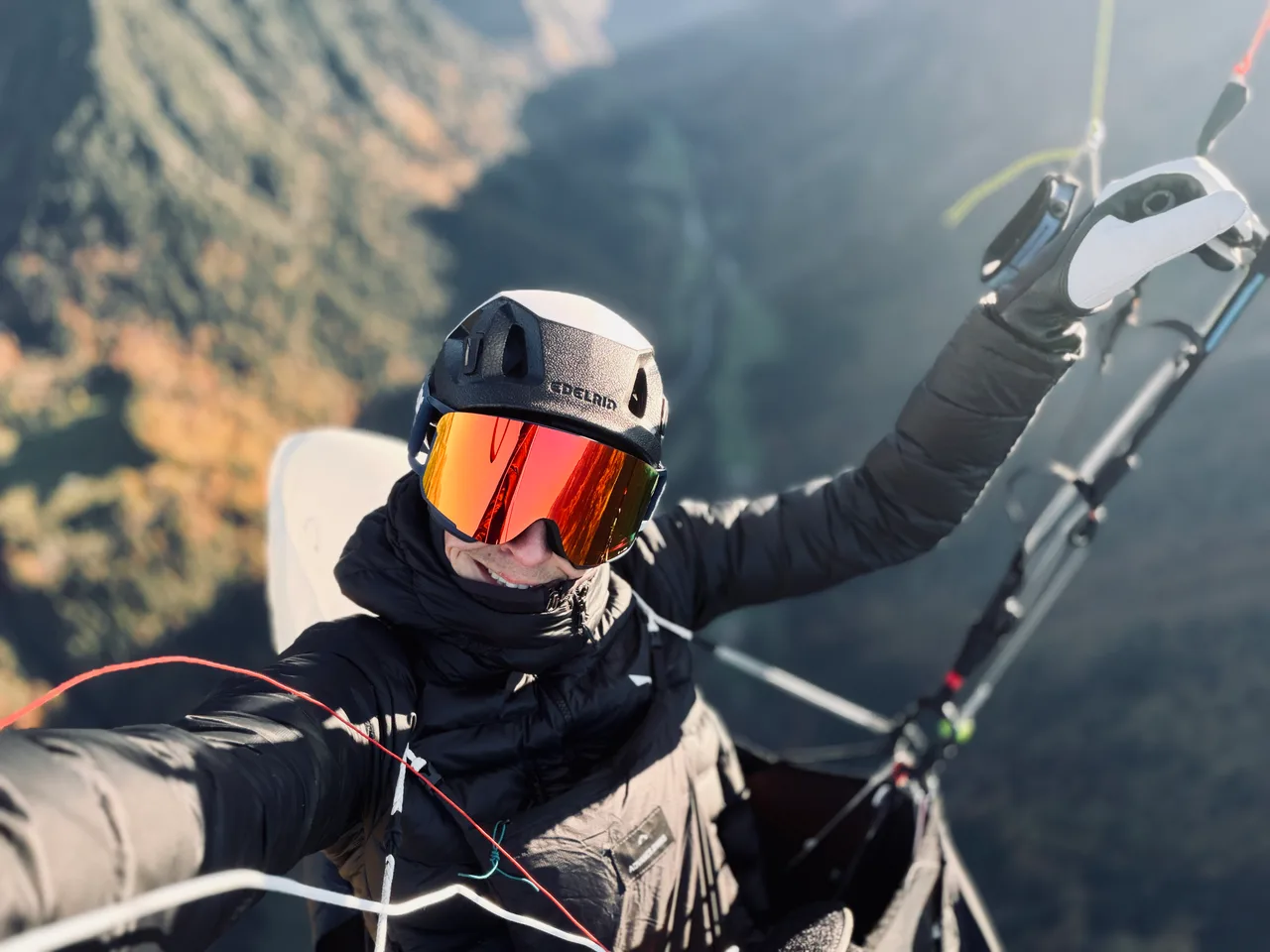 Gleitschirmpilot mit Helm und verspiegelter Brille im Flug vor einem bewaldeten Berghang