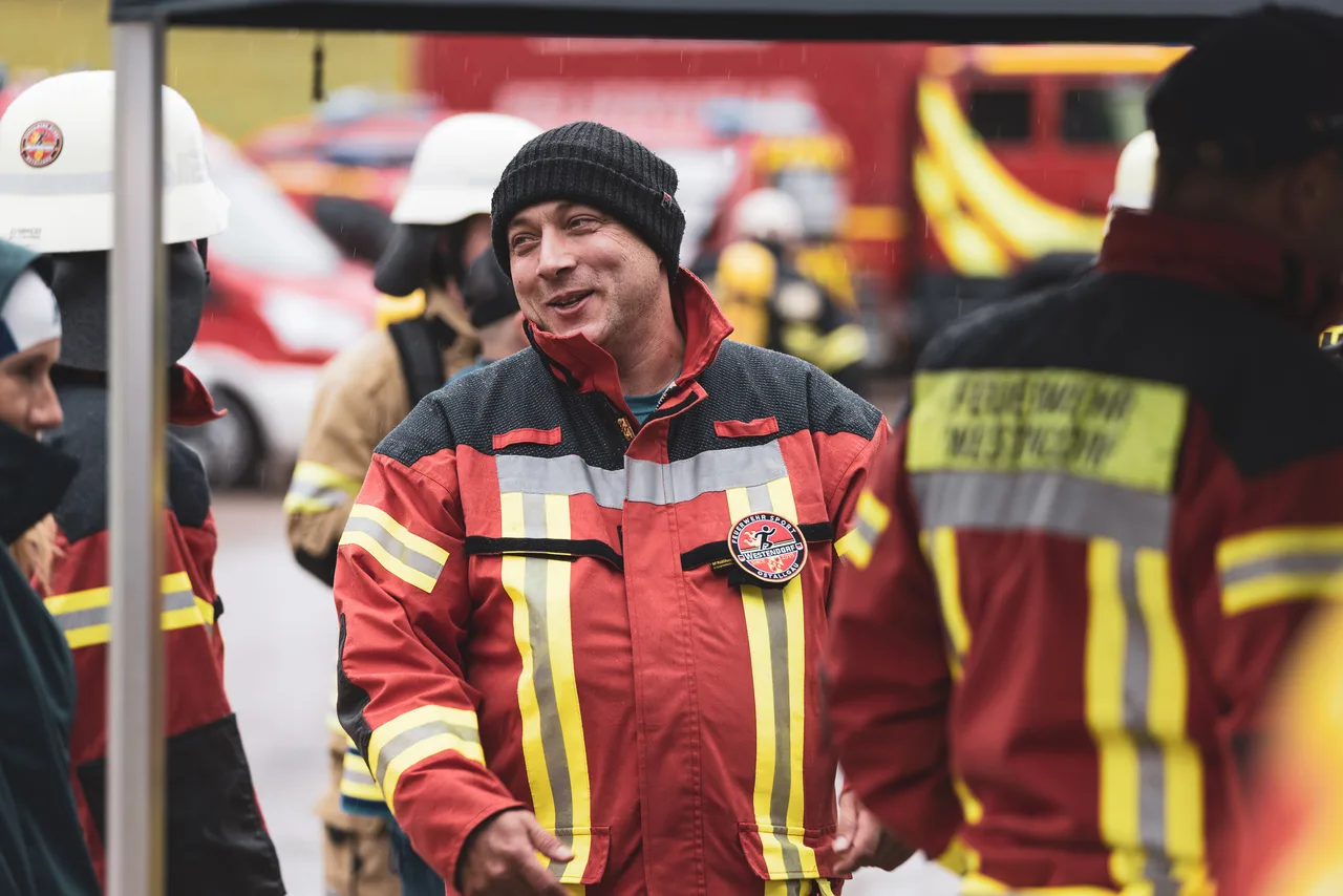 Feuerwehrmann in roter Schutzjacke und schwarzer Mütze im Gespräch mit anderen Teilnehmern im Regen.