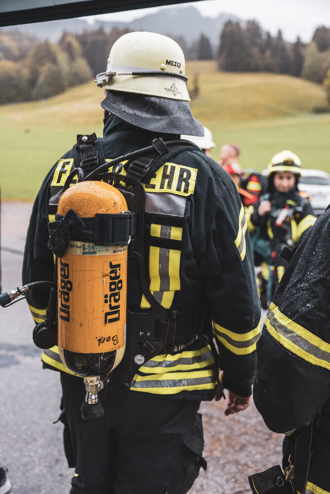 Regentropfen auf Helm und gelber Atemluftflasche eines Feuerwehrteilnehmers, von hinten gesehen.