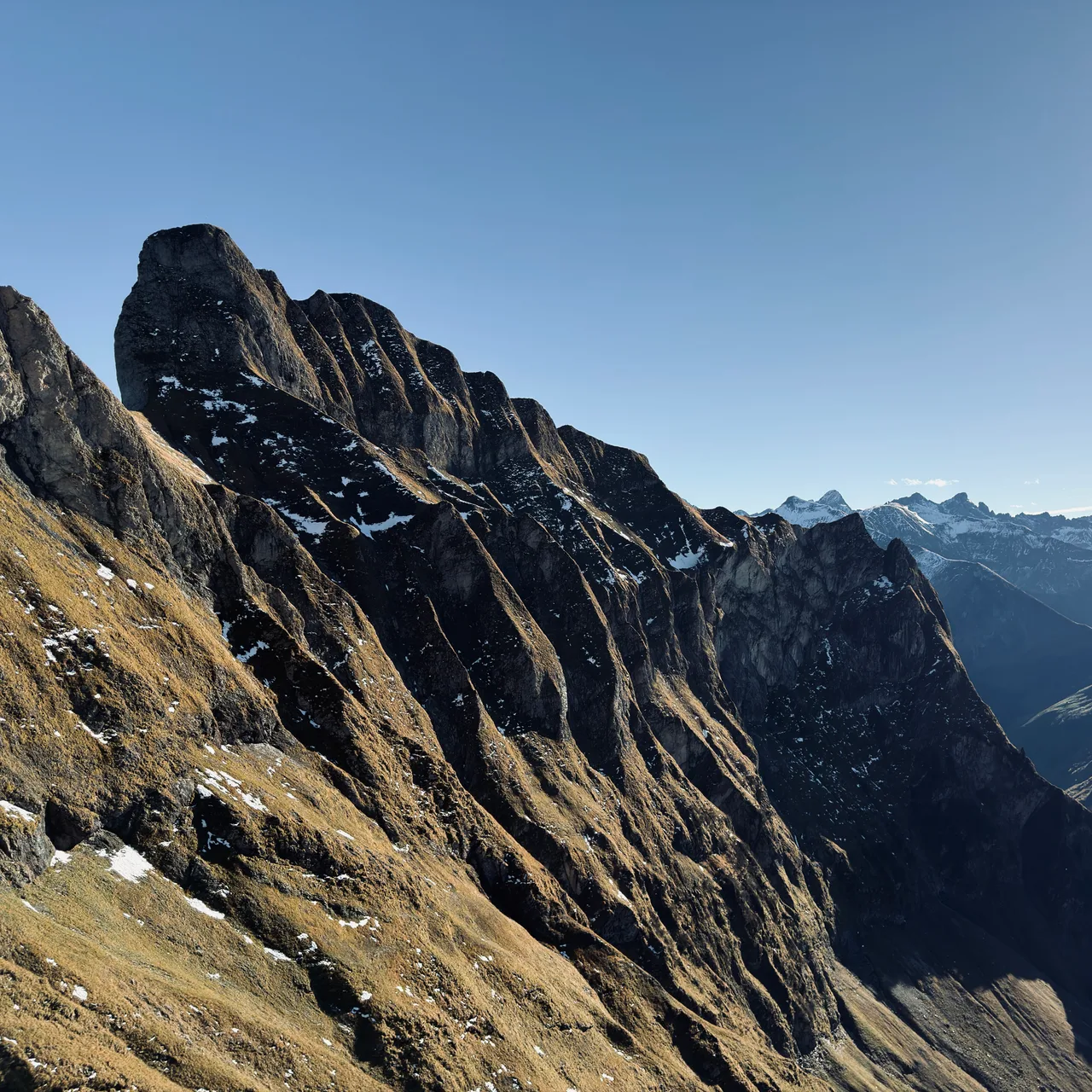 Steile Fels- und Grasflanken des Schnecks mit Schneeresten vor schneebedeckten Gipfeln unter blauem Himmel