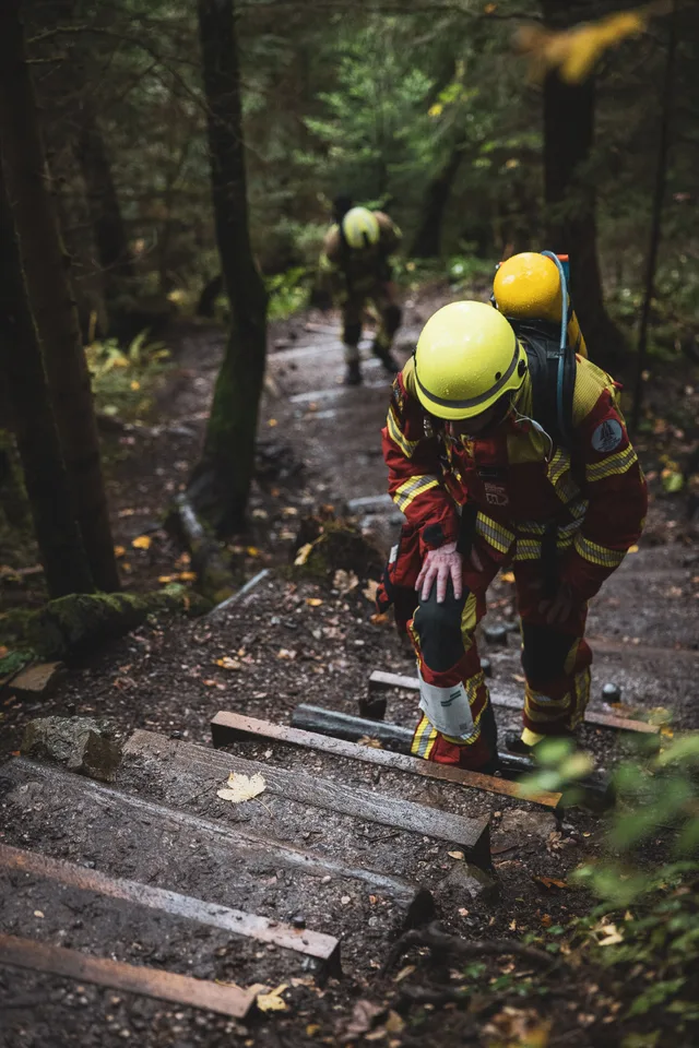Teilnehmer in roter Einsatzkleidung beugt sich beim Aufstieg über die Waldstufen nach vorn.
