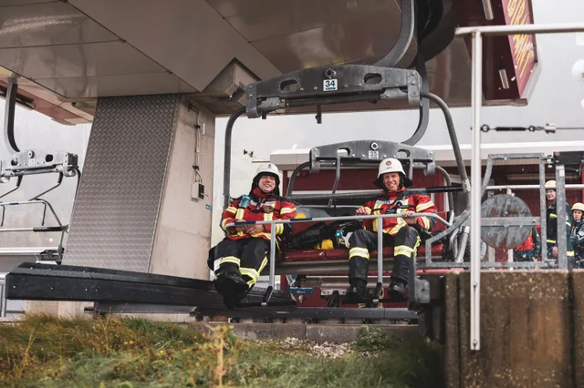 Zwei Teilnehmer in roter Einsatzkleidung fahren mit dem Sessellift aus der Station.