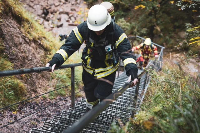 Teilnehmer in schwarzer Einsatzkleidung und weißem Helm steigt die Waldtreppe hinauf.