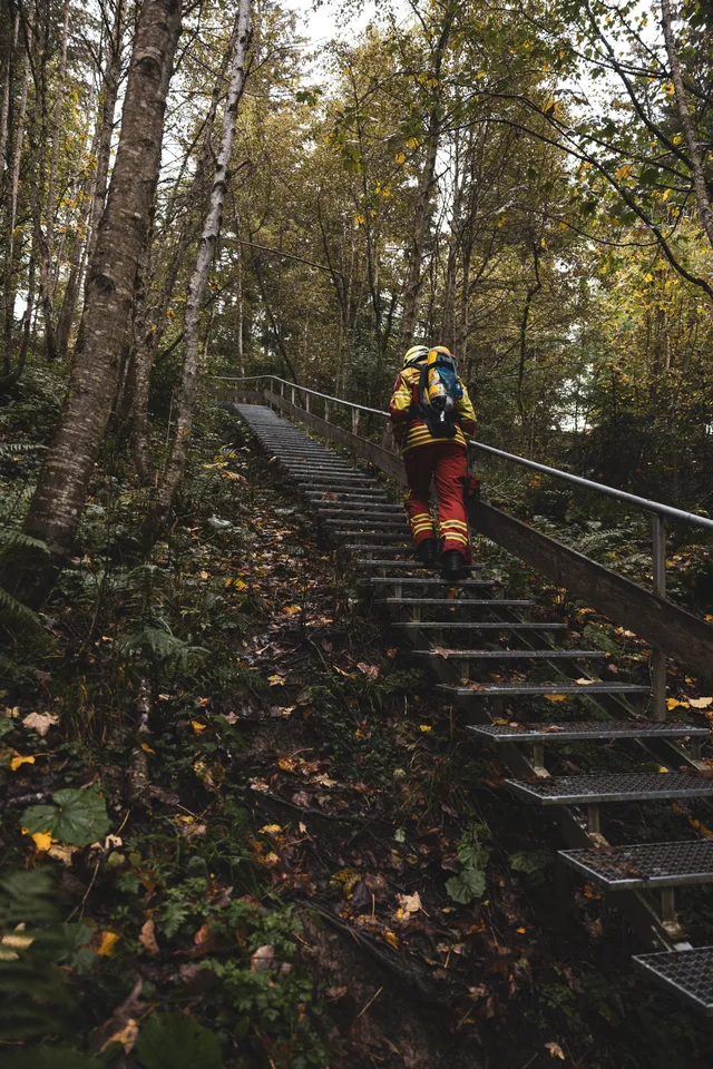 Teilnehmer in roter Schutzkleidung steigt mit gelber Atemluftflasche eine lange Waldtreppe hinauf.