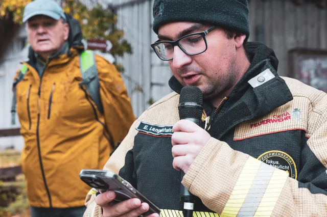 Teilnehmer mit Strickmütze spricht ins Mikrofon und hält ein Mobiltelefon.