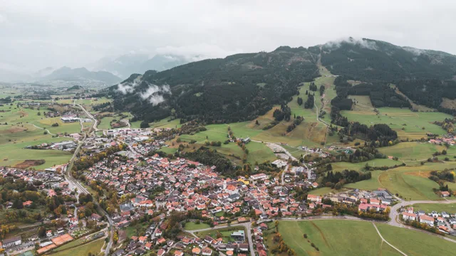Luftaufnahme von Nesselwang und den bewaldeten Hängen der Alpspitze unter tiefen Wolken.