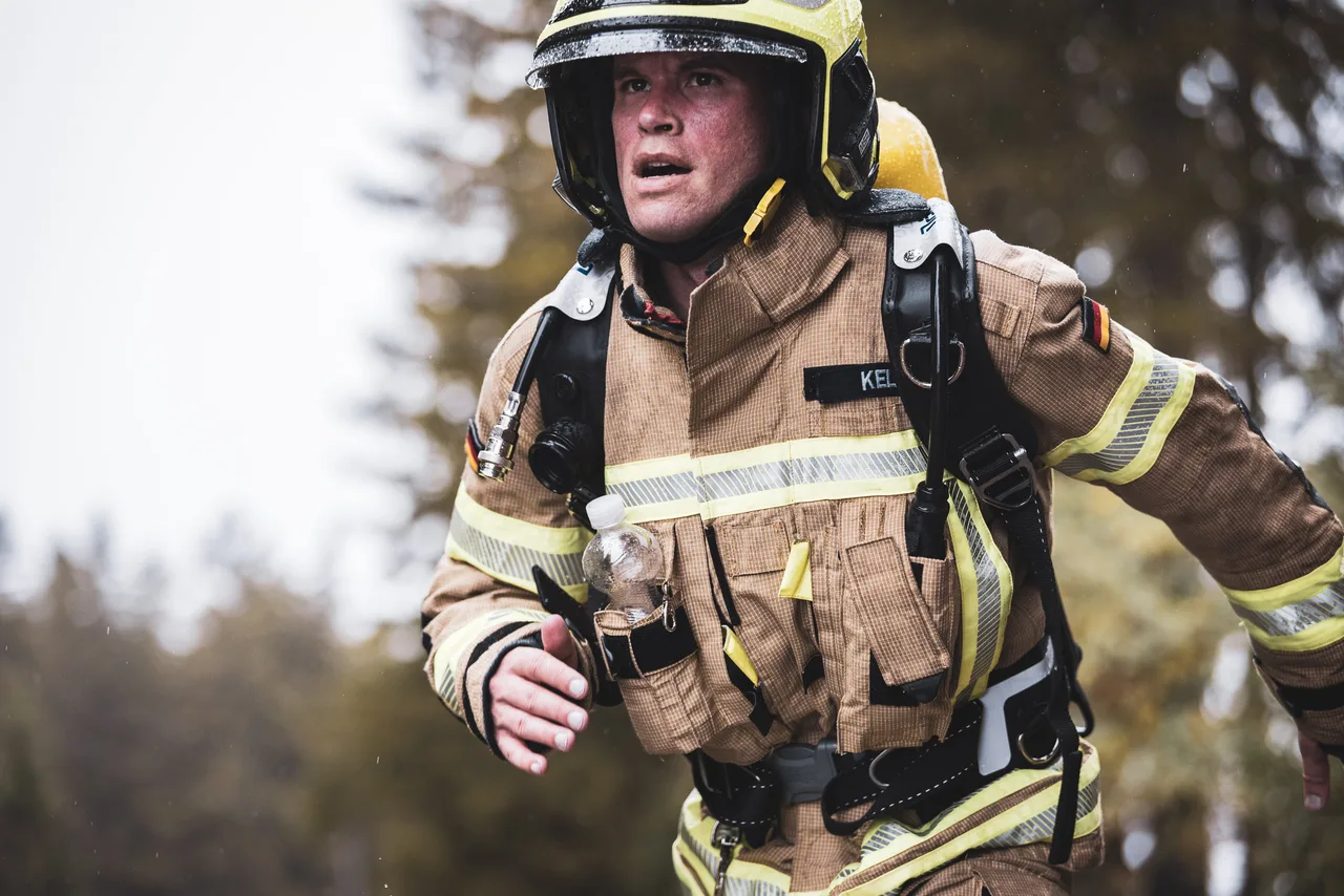 Feuerwehrmann mit gelbem Helm und Atemschutzgerät steigt mit geöffnetem Mund im Regen bergauf.