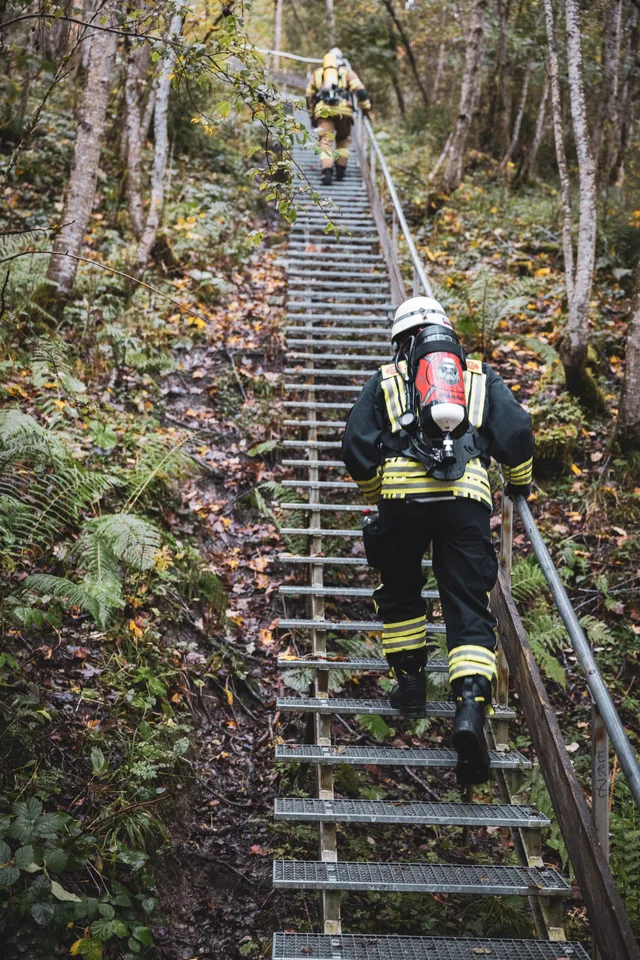 Teilnehmer in dunkler Einsatzkleidung auf einer steilen Metalltreppe im Wald.